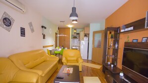 A living room with a yellow sofa, a coffee table, a TV, and a kitchen in the background.