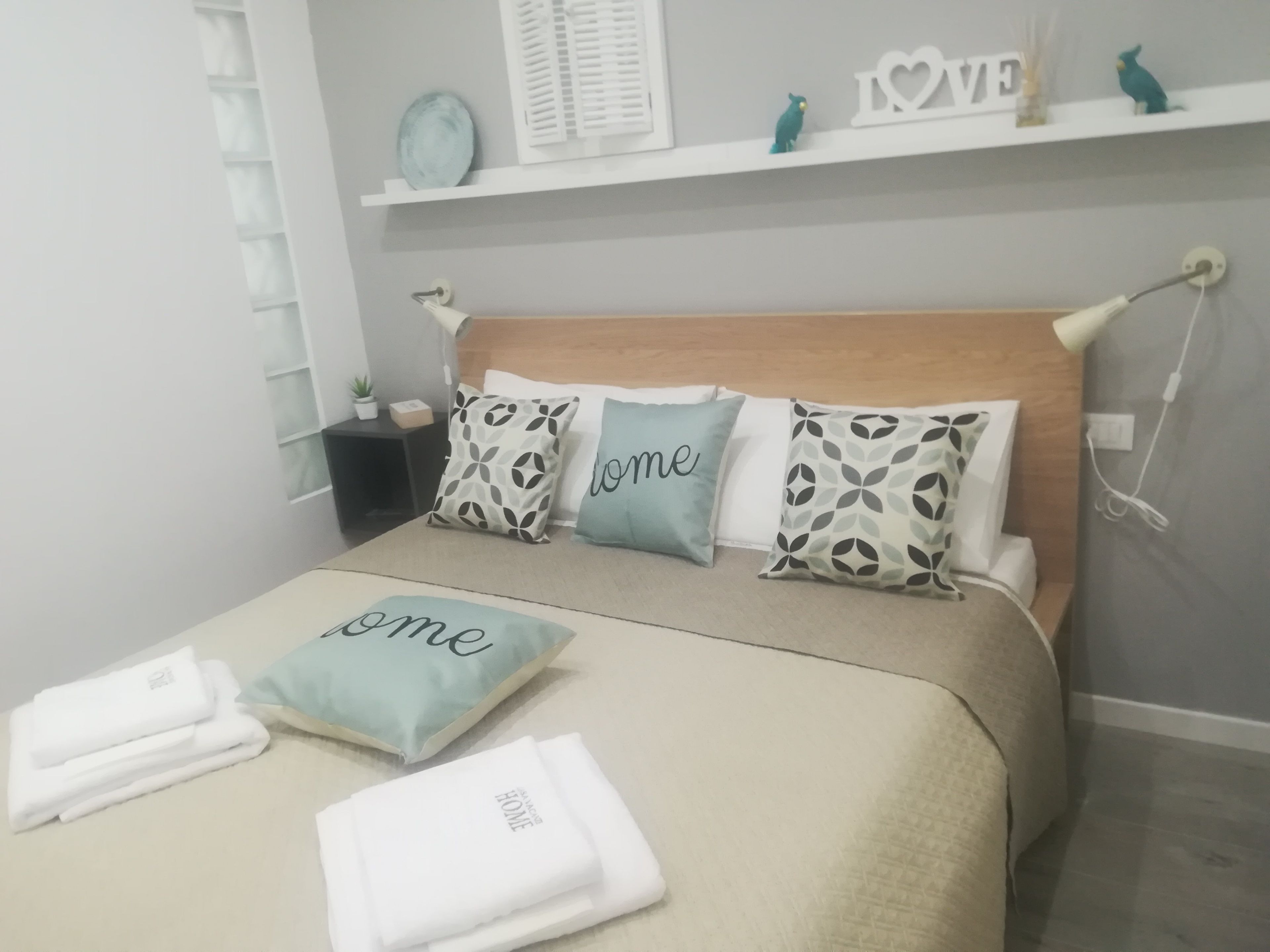 Una cama bien hecha con almohadas que tienen la palabra 'home', una mesita de noche con una lámpara y una repisa sobre la cama con objetos decorativos.