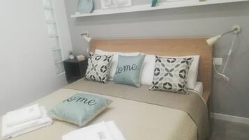 Una cama bien hecha con almohadas que tienen la palabra 'home', una mesita de noche con una lámpara y una repisa sobre la cama con objetos decorativos.