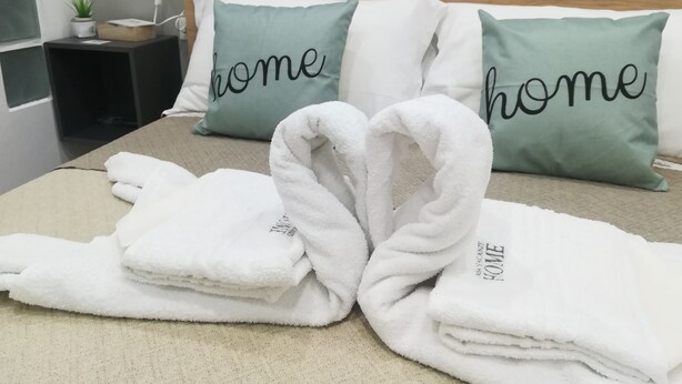 Ein ordentlich bezogenes Bett mit zwei Kissen, eines mit dem Aufdruck "home", und einem kleinen Nachttisch mit einer Lampe und einer Pflanze.