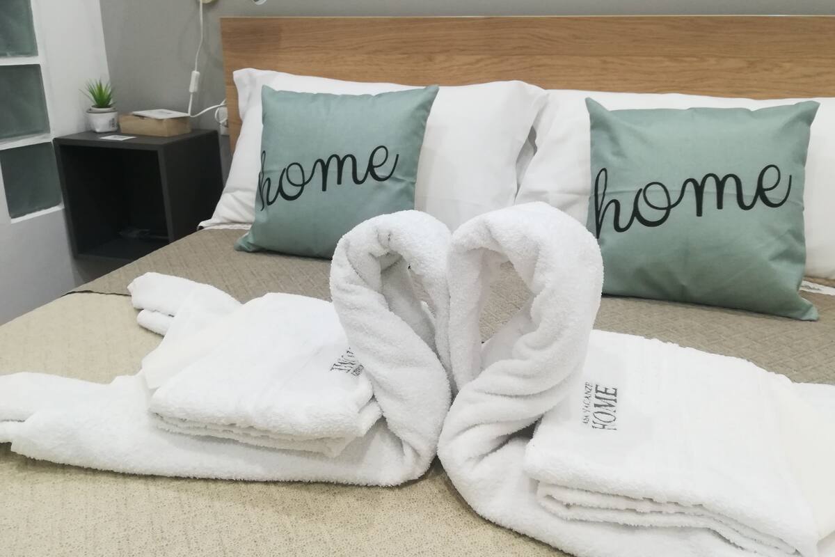 A neatly made bed with two pillows, one with "home" written on it, and a small bedside table with a lamp and a plant.