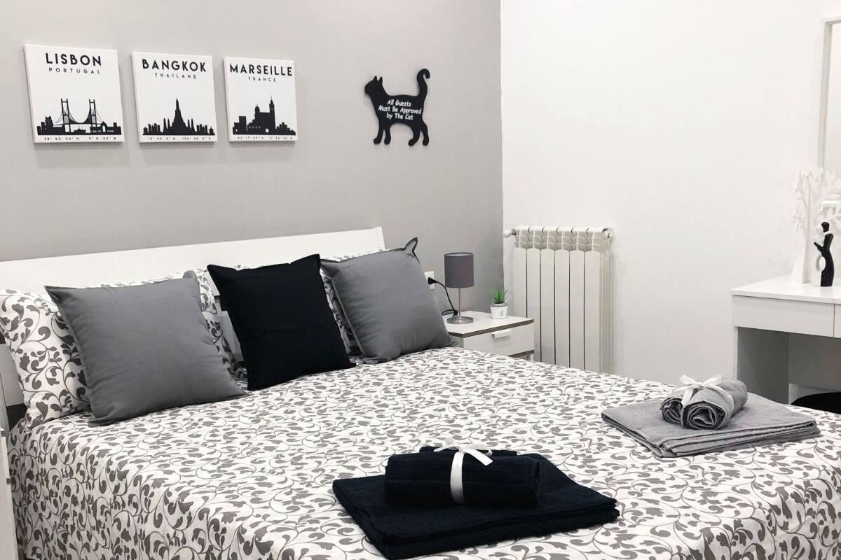 A neatly made bed with a patterned bedspread, a black and white cat silhouette on the wall, and three cityscape pictures.