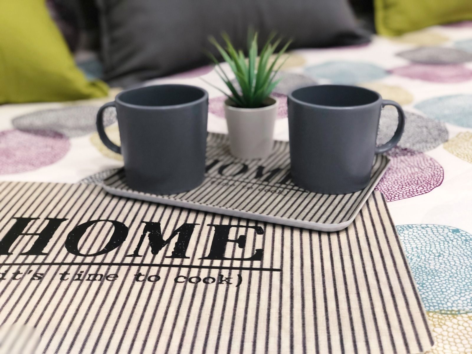 Eine Küchenecke mit zwei schwarzen Tassen auf einem Tablett, einer kleinen Topfpflanze und einer Unterlage mit dem Wort 'HOME' und dem Spruch 'Zeit zu kochen'.