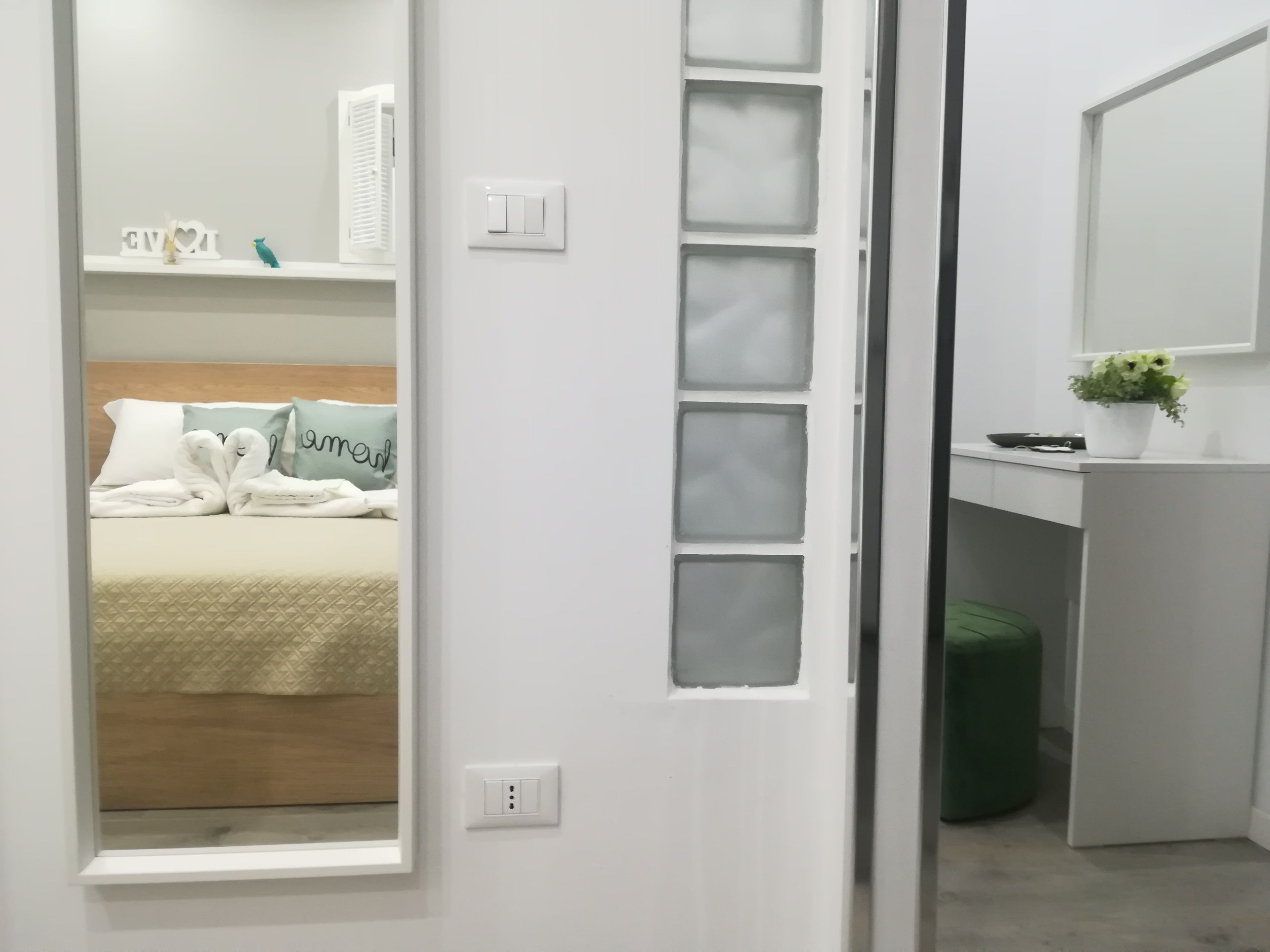 A hotel room with a bed, a bedside table, a mirror, and a green trash bin.