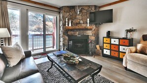 A cozy living room with a stone fireplace, a flat-screen TV mounted on the wall, a leather sofa, a coffee table with decorative items, and a colorful cabinet.