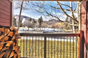 A wooden deck with a railing, a stack of firewood, and a view of a snowy landscape with trees and a road.