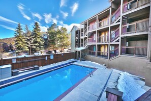 A swimming pool with a wooden deck, a fenced area, and a multi-story building with balconies.