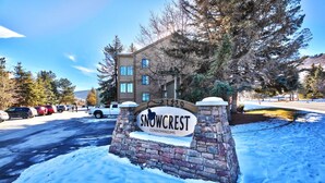 A sign for Snowcrest Condominiums with a stone base and a wooden top, surrounded by snow and parked cars.