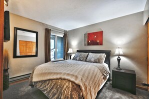A bedroom with a bed, two bedside lamps, a window with curtains, a framed picture on the wall, and a painting above the bed.