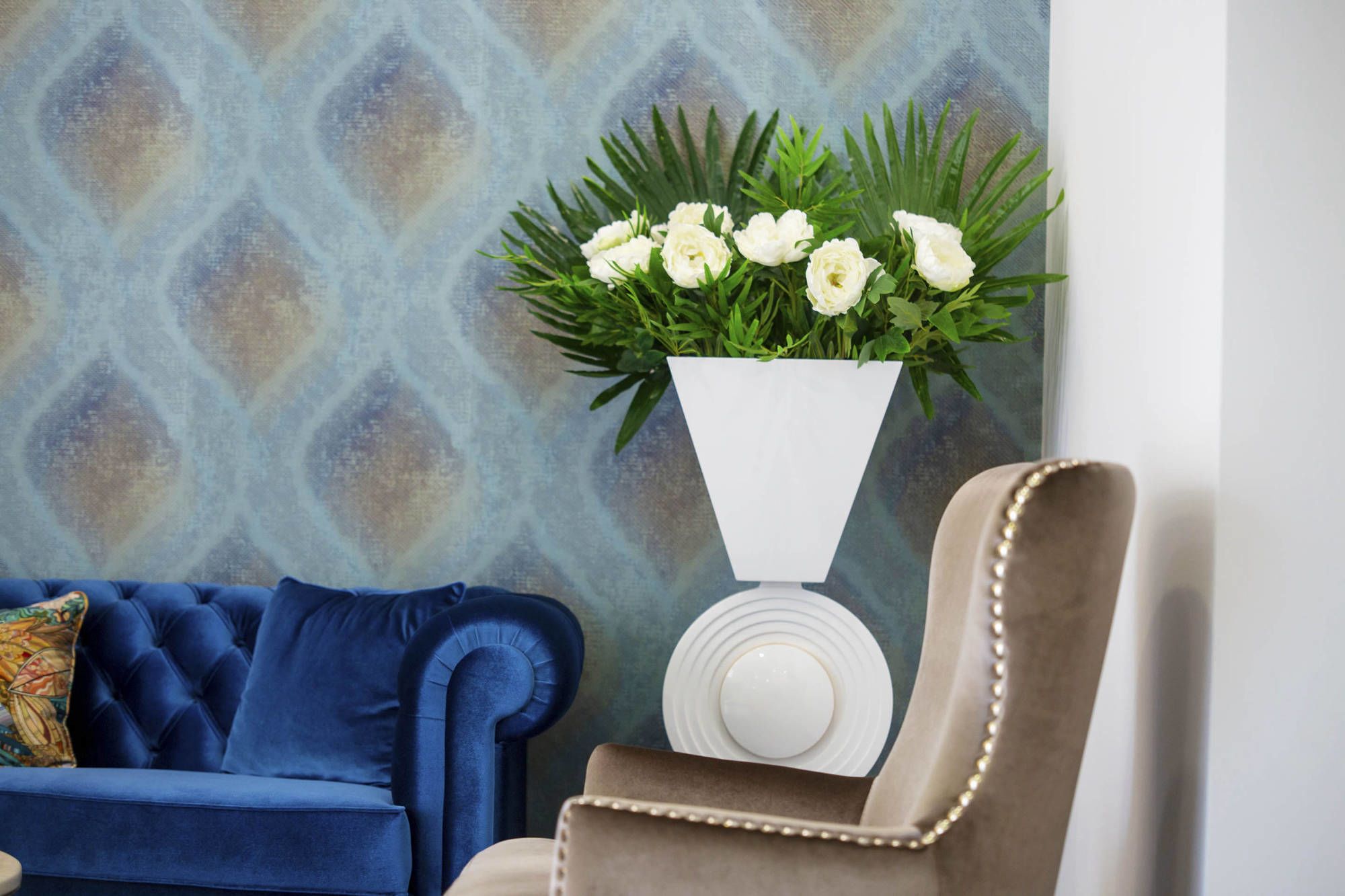 A blue sofa with a patterned cushion, a white vase with white flowers, and a beige armchair with a decorative backrest.