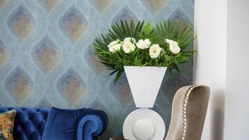 A blue sofa with a patterned cushion, a white vase with white flowers, and a beige armchair with a decorative backrest.