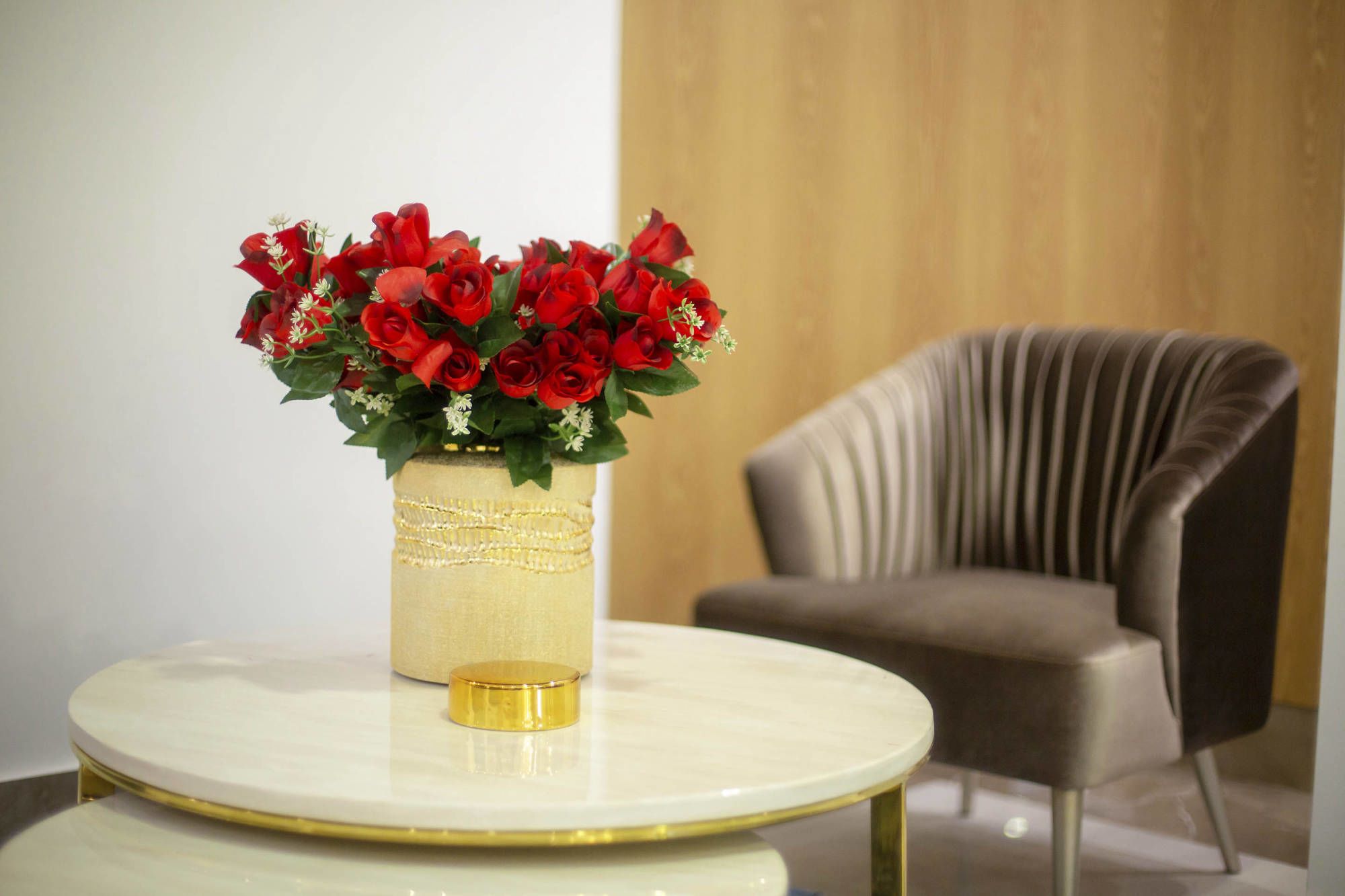 A vase with red roses on a round table in a room with a modern chair.