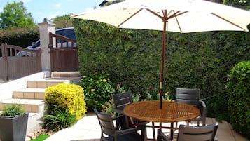 Een terras met een ronde houten tafel en vier stoelen, een parasol en een haag.
