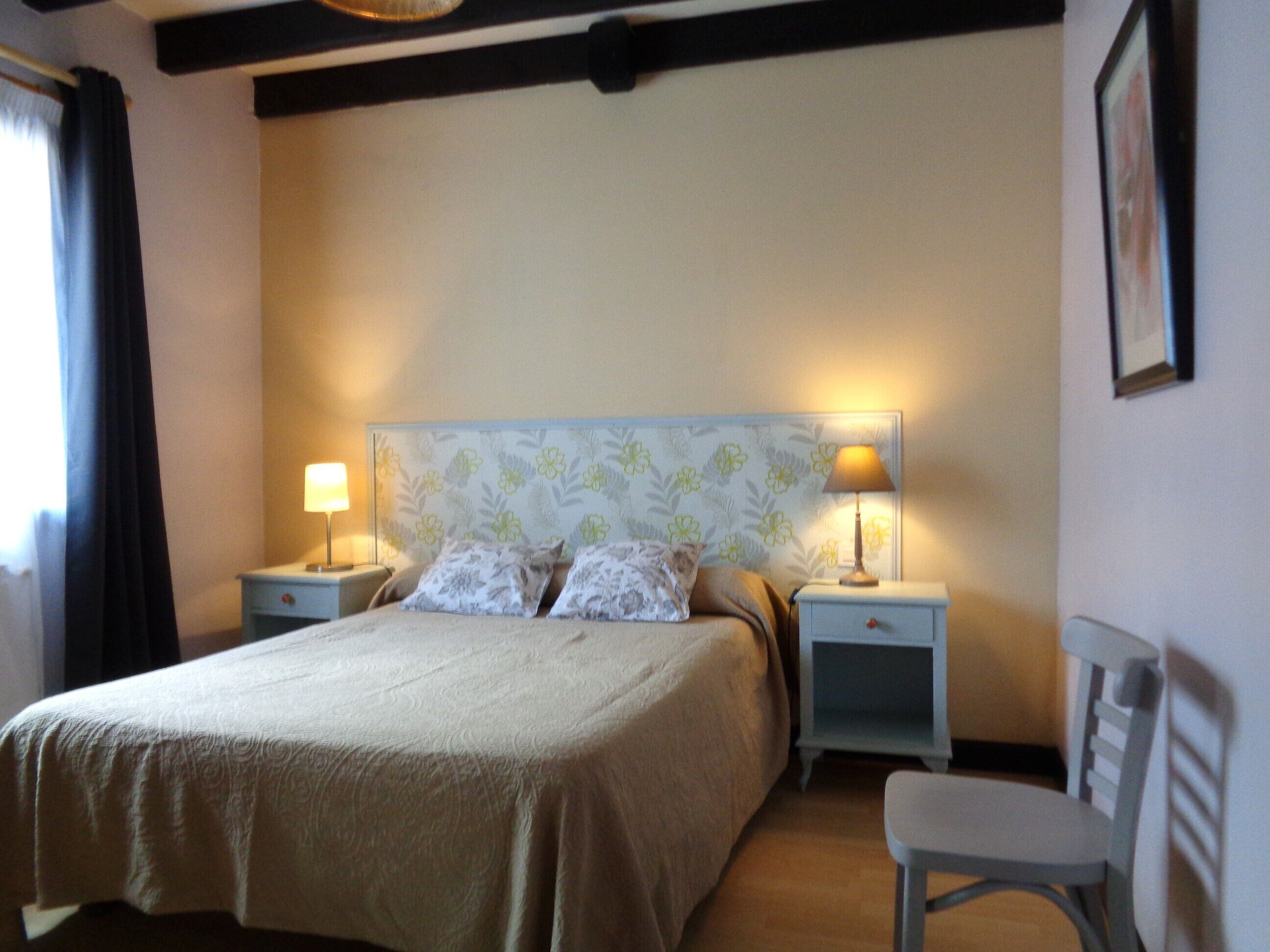 Un dormitorio ordenado con una cama, dos mesitas de noche con lámparas, una silla y un cuadro en la pared.