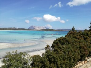 A coastal landscape with clear turquoise waters, a sandy beach, and a mountainous backdrop.
