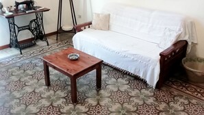 Ein Wohnzimmer mit einem gemusterten Boden, einem weißen Sofa, einem Holztisch und einer Nähmaschine, sowie einem gerahmten Gemälde an der Wand.