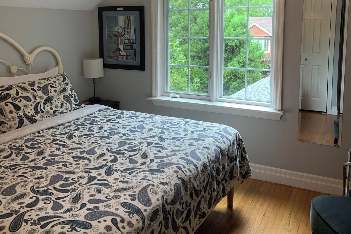 A bedroom with a bed, a ceiling fan, a window with a view of greenery, and a door.