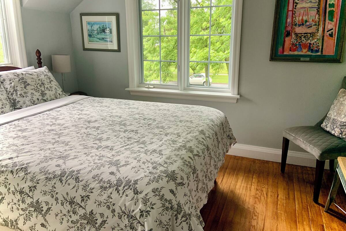 A bedroom with a large bed, a window with a view of greenery, a chair, and a framed picture on the wall.