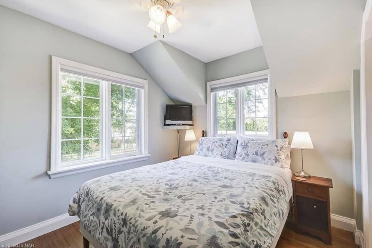 A bedroom with a large bed, two windows, a ceiling fan, and a TV mounted on the wall.