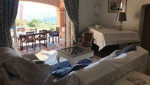 A room with a sofa, a dining table with chairs, a small table with a lamp, and a view through an archway to an outdoor seating area. - Villa with swimming pool in the Gulf of SAINT-TROPEZ (La Croix-Valmer)