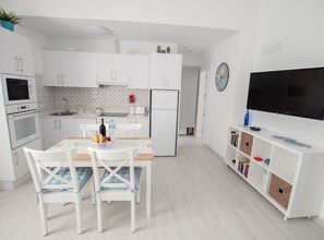 Eine moderne Küche mit weißen Schränken, einem Esstisch mit Stühlen, einem Kühlschrank, einer Spüle und einem Fernseher auf einem weißen Schrank.