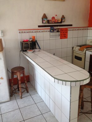 A kitchen with a white countertop, a wooden stool, a microwave, a stove, and a shelf with kitchen items. - Hostal Brisas y Olas - Hostel (San Juan del Sur)