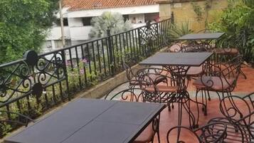 Un patio exterior con mesas y sillas, rodeado por una valla de hierro forjado negro, con vistas a una colina con edificios.