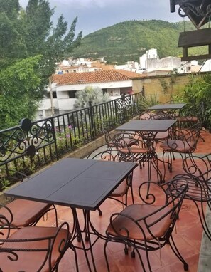 Een terras buiten met tafels en stoelen, omgeven door een zwart ijzeren hek, met uitzicht op een heuvel met gebouwen.