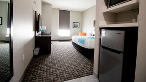 A hotel room with a bed, a refrigerator, a microwave, a desk with a lamp, and a window with blinds. - Hotel Pryor (Pryor)