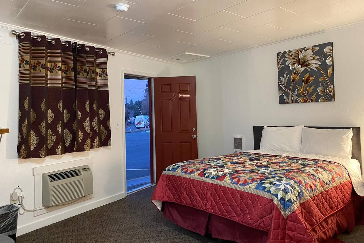 Ett hotellrum med en säng, ett fönster med gardiner, en dörr, en tavla på väggen och en luftkonditioneringsanläggning.