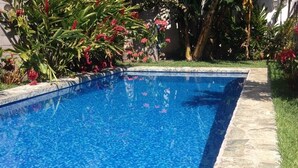 Piscina retangular com borda de mosaico azul, rodeada por um deck de pedra e vegetação exuberante.