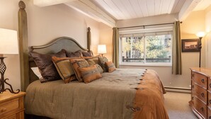 Een slaapkamer met een groot bed, een houten dressoir, een nachtkastje, een raam met gordijnen en een lamp.