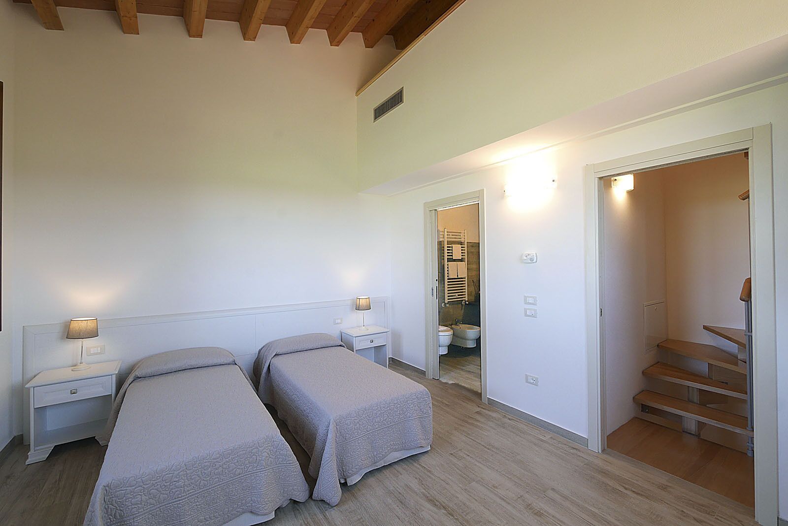 Un dormitorio con dos camas individuales, una mesita de noche con lámpara, techo de madera y una escalera que conduce a un piso superior.