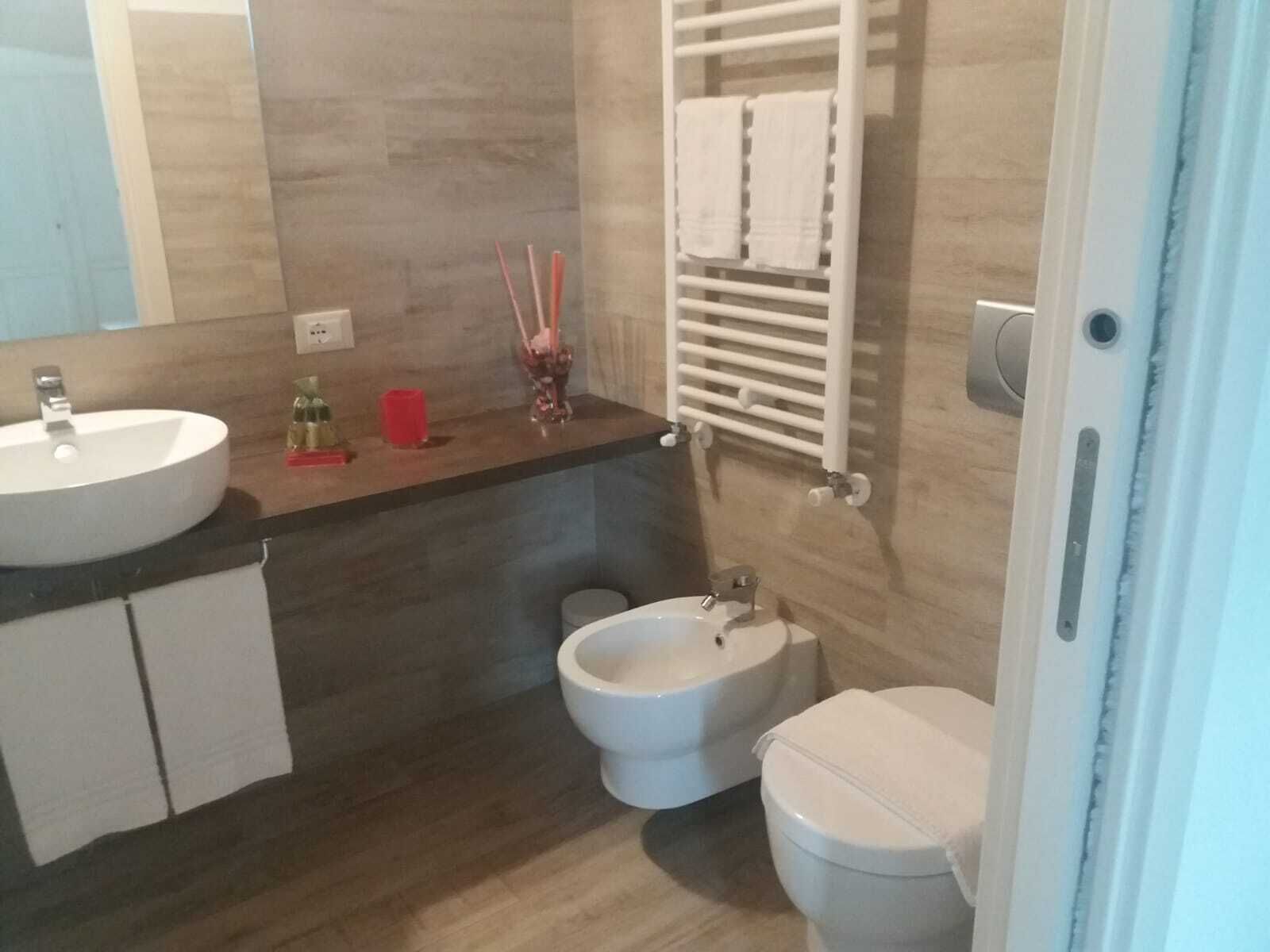 A modern bathroom with a sink, bidet, and toilet. There is a towel rack with towels, a mirror, and a small shelf with toiletries.