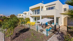 A modern two-story building with a flat roof, large windows, and a balcony. There are yellow chairs and a white umbrella outside. - Ocean Villas-Villa Sol (Guia)
