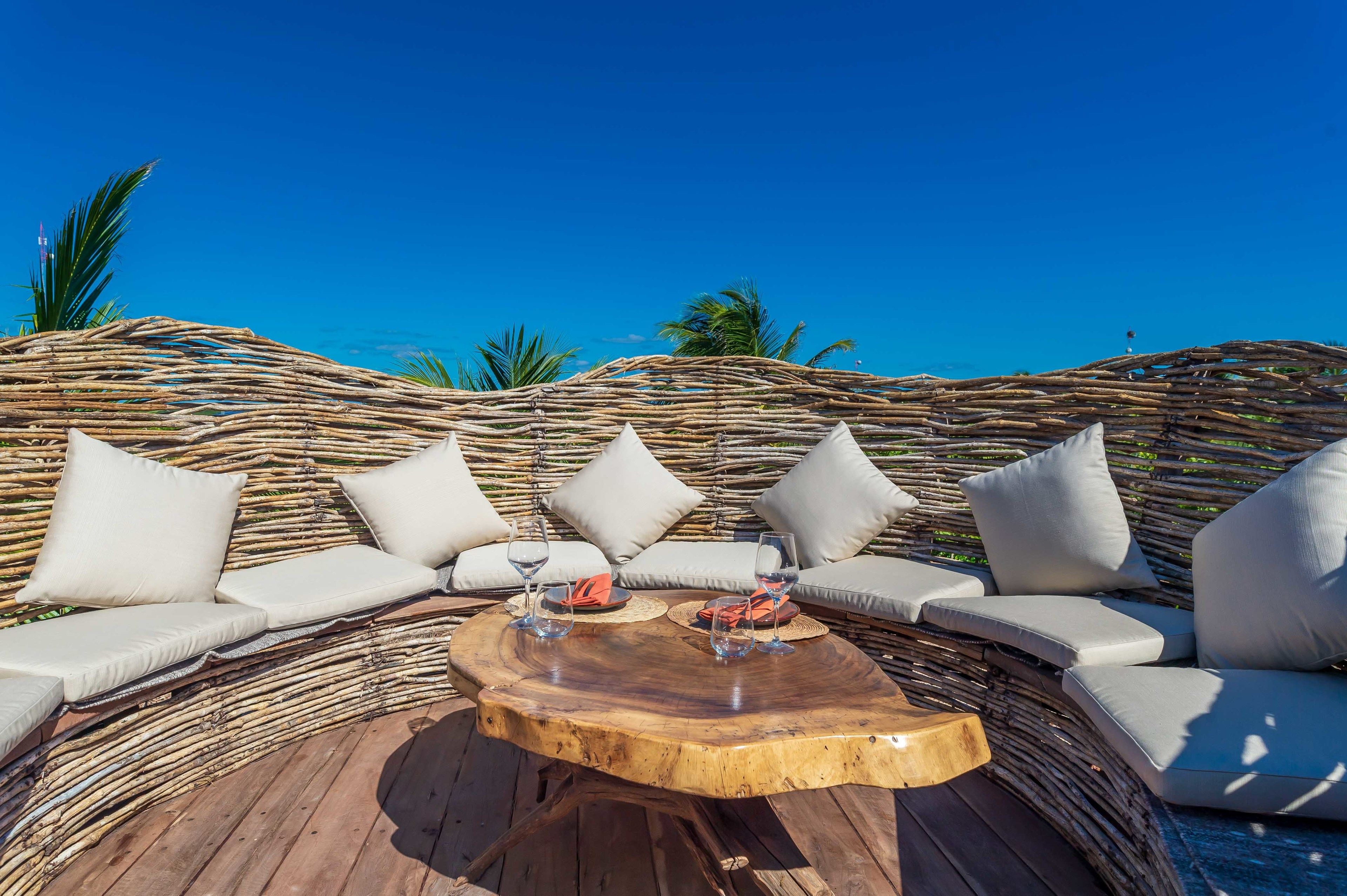 A wooden deck with wicker furniture and cushions, a round wooden table, and a clear blue sky.