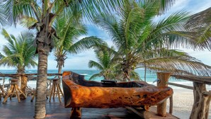 Una escultura de barco de madera sobre una cubierta de madera, con palmeras y vista a la playa.