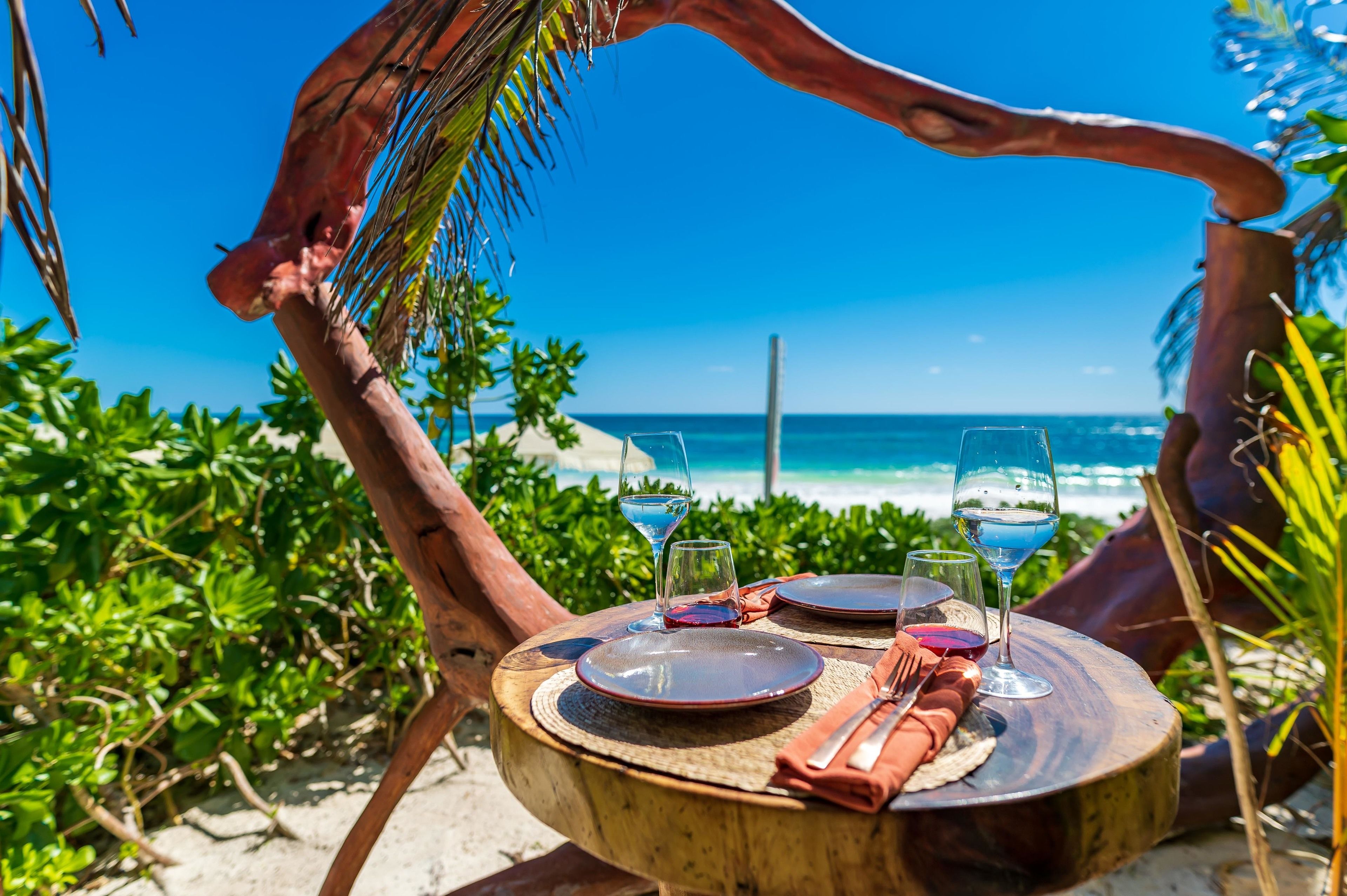 Una mesa de madera con platos, vasos y cubiertos, colocada al aire libre con vista al océano.