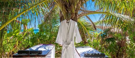 A white robe hanging from a palm tree, two massage tables with black mats, and a tropical setting with dense foliage.
