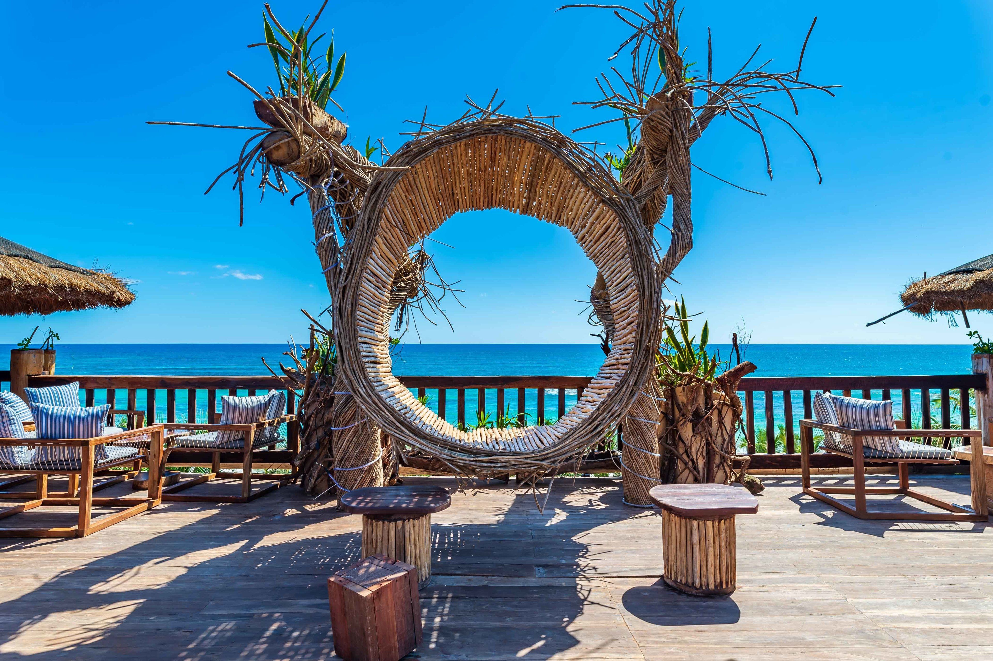 Estructura circular tejida con sillas y mesas sobre una terraza de madera con vista al océano.