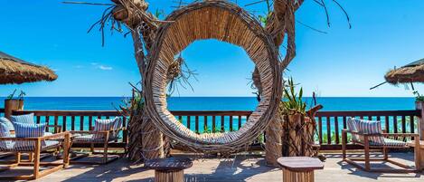 A circular woven structure with chairs and tables on a wooden deck overlooking the ocean.
