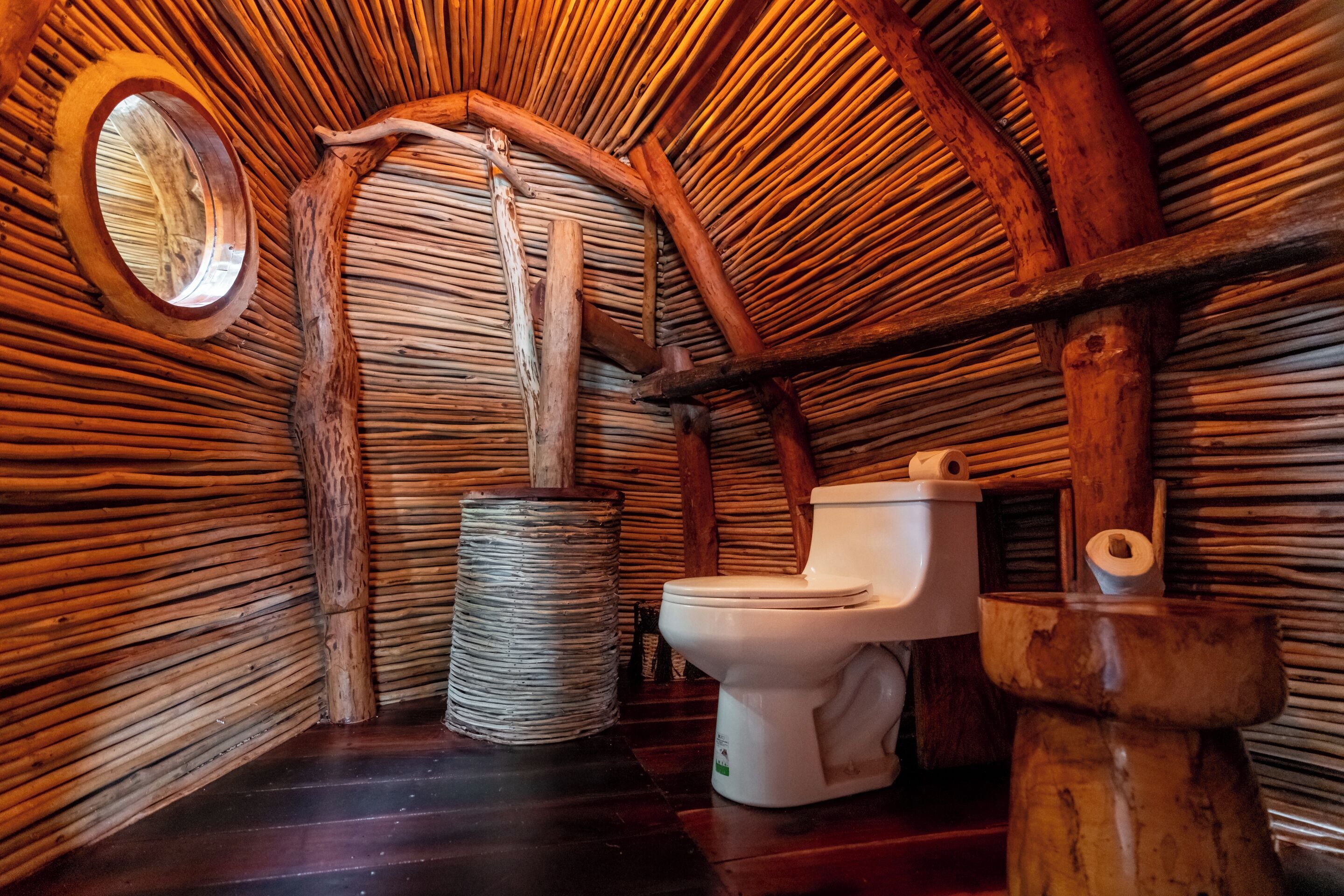 Une salle de bain rustique avec un plafond en chaume, des toilettes en bois et un miroir rond.