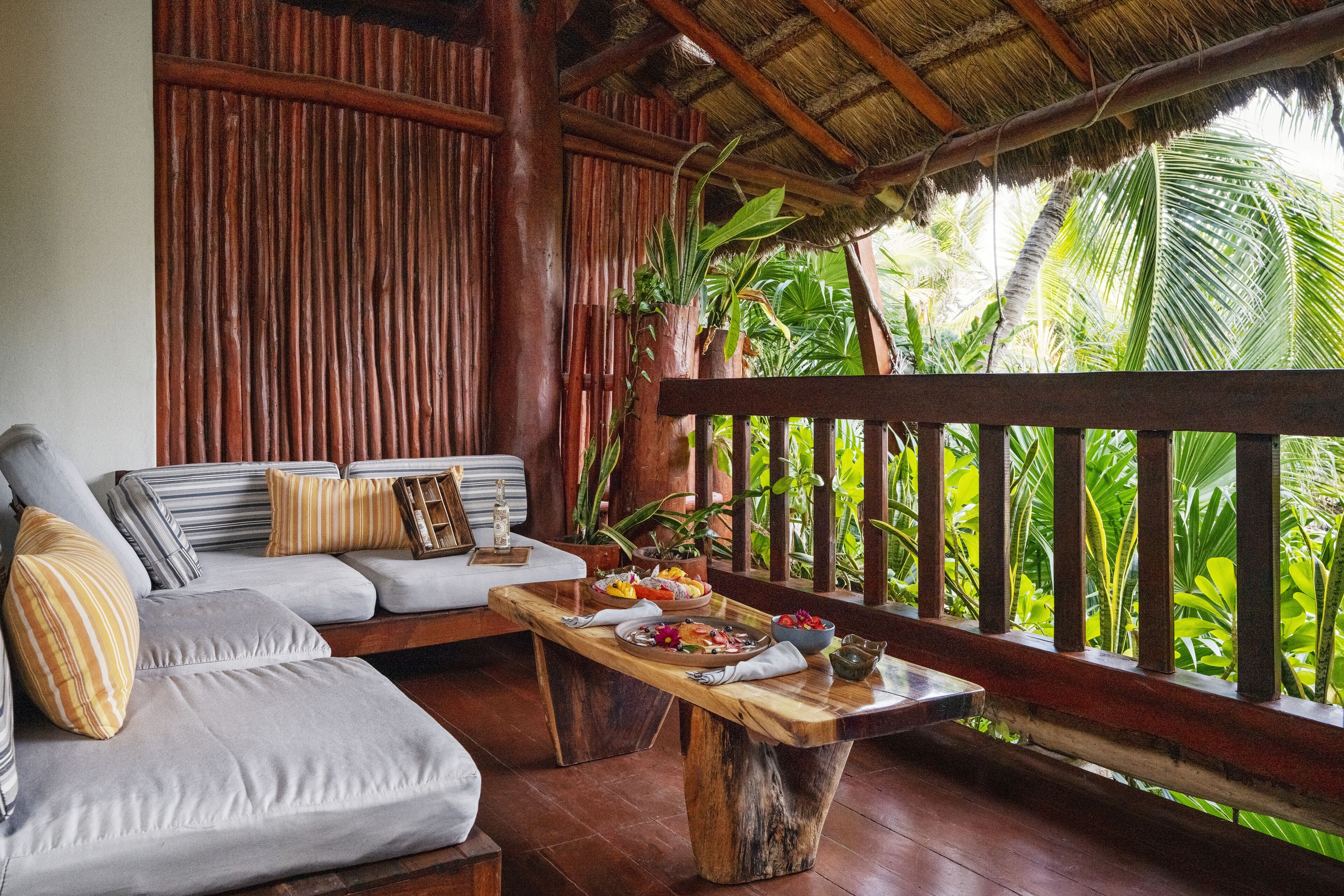 A wooden deck with a sofa and a dining table set with food, surrounded by lush greenery.