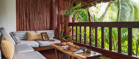 A wooden deck with a sofa and a dining table set with food, surrounded by lush greenery.