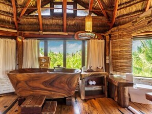 A wooden bathtub in a room with a thatched roof, wooden benches, and a view of greenery outside.