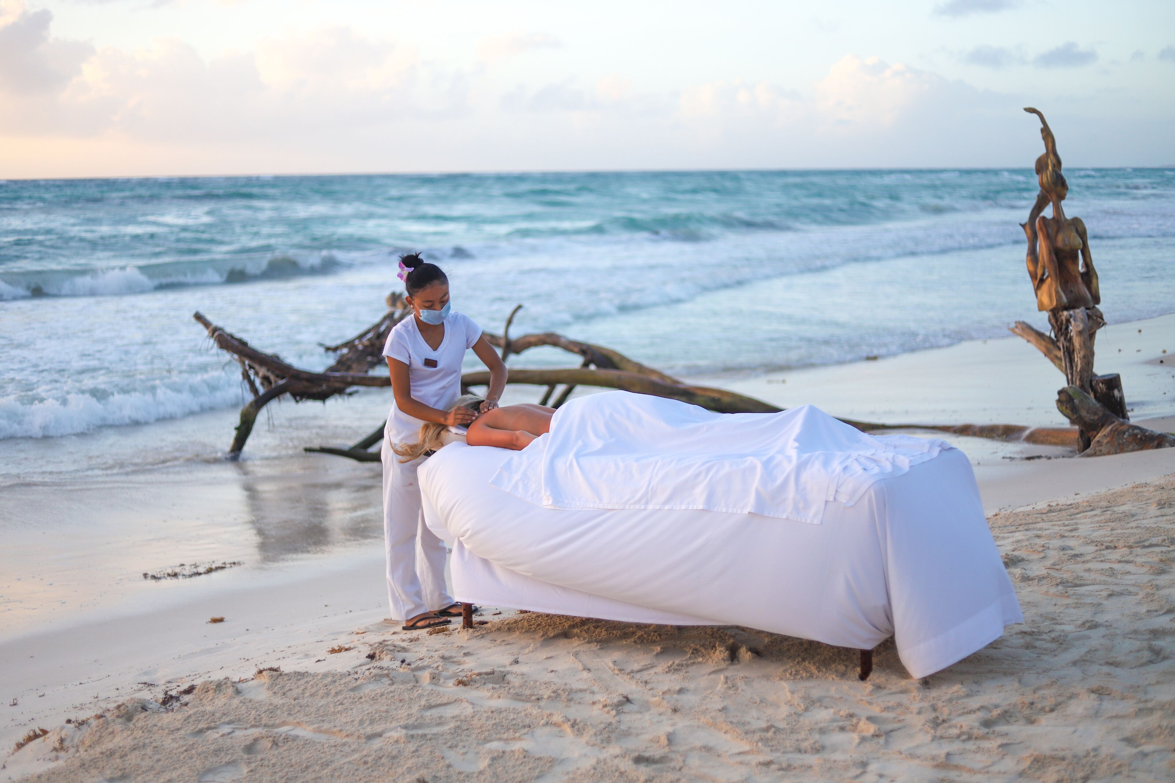 A beach scene with a person lying on a white-covered bed, another person attending to them, and driftwood in the background.