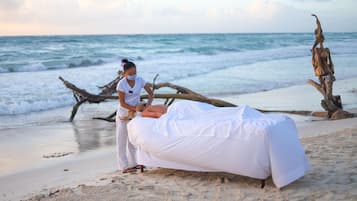 A beach scene with a person lying on a white-covered bed, another person attending to them, and driftwood in the background.