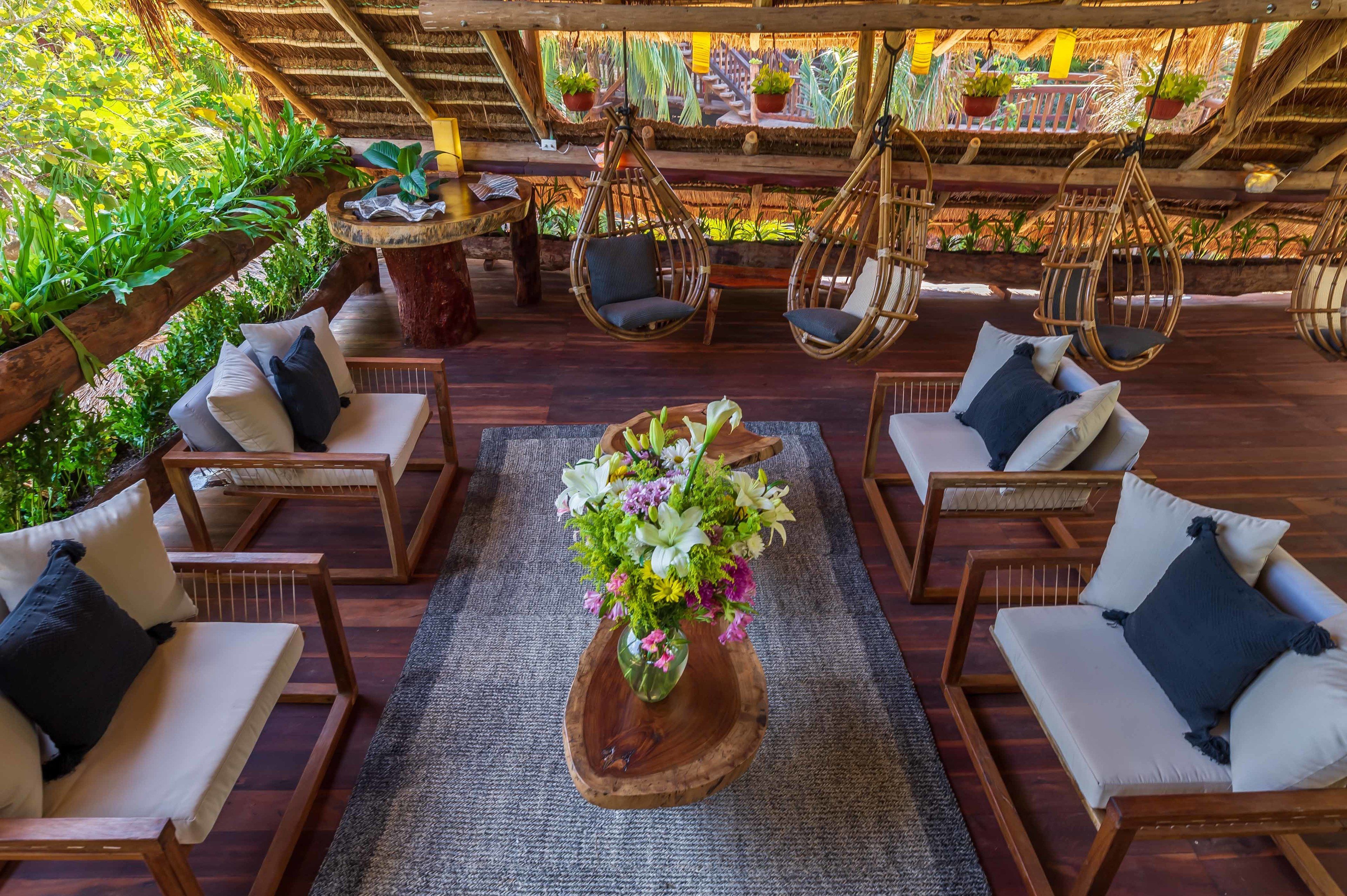 A wooden deck with wicker chairs and sofas, a central wooden table with a floral arrangement, and hanging chairs.