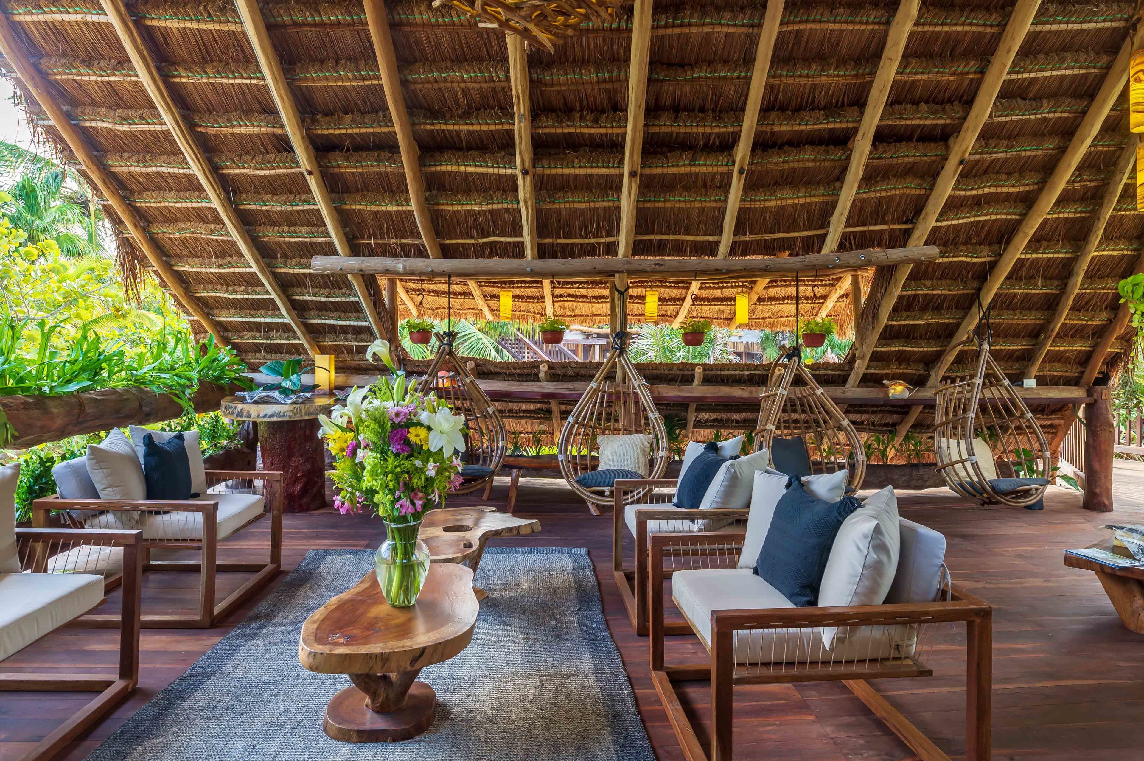 An outdoor seating area with wooden furniture, hanging chairs, and potted plants under a thatched roof.