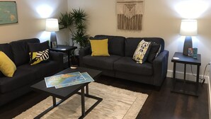 A living room with a dark grey sofa, a coffee table with a map, two side tables with lamps, a plant, and a wall map. - DaLight - A beautiful home in a quiet neighborhood (Kailua)